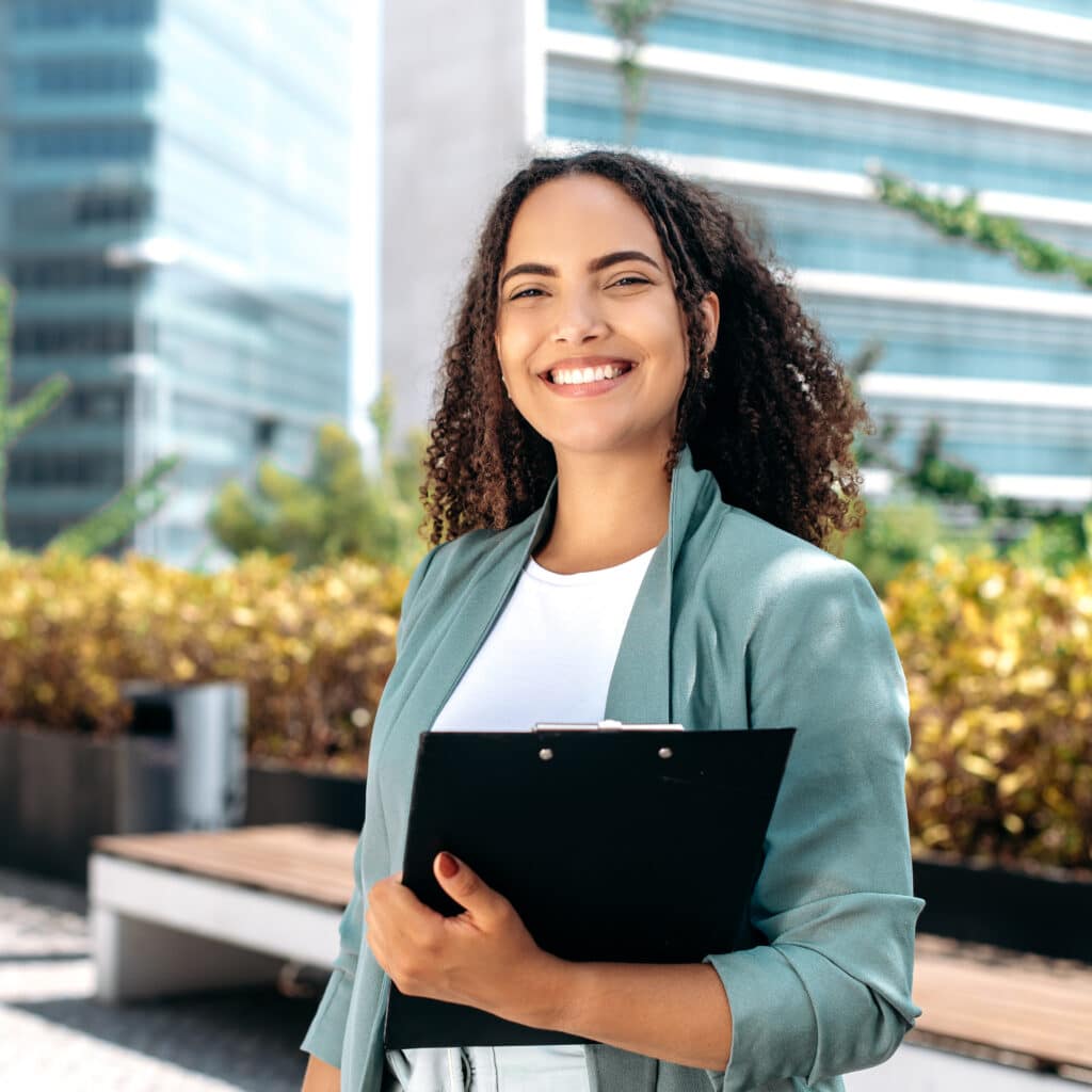 Jeune professionnelle souriante avec presse-papiers en extérieur Jeune femme souriante aux cheveux bouclés, vêtue d'une veste bleu-vert, tenant un presse-papiers noir à l'extérieur d'un immeuble de bureaux moderne.